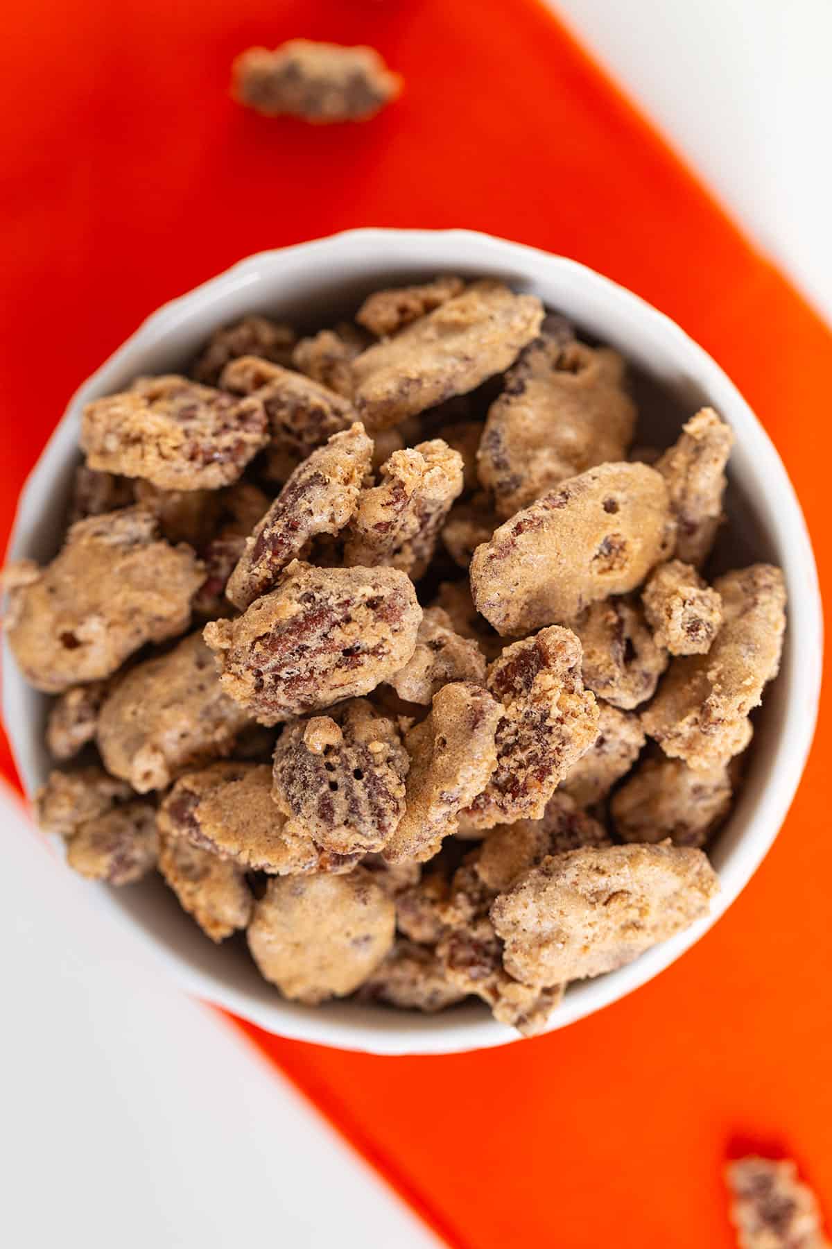 A white bowl filled with candied pecans sits on an orange napkin, showcasing the rich glaze and crunch of these irresistible candied pecans, viewed from above.