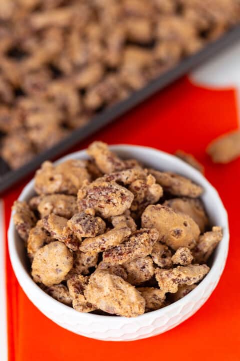 A white bowl filled with delicious candied pecans sits on an orange napkin, with a tray of extra candied pecans in the background.
