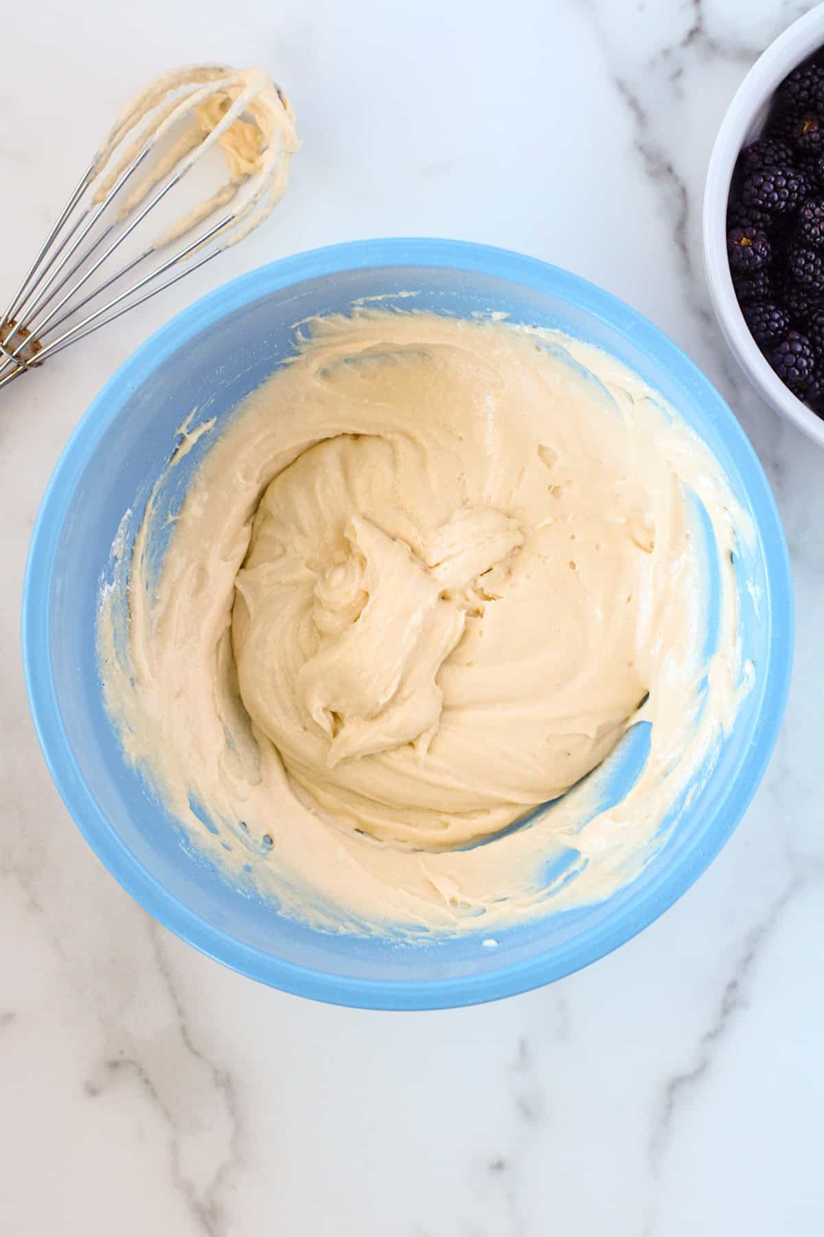 A blue bowl filled with cake batter sits on a marble countertop next to a whisk and a white bowl of blackberries, ready to be transformed into a delicious blackberry cobbler.
