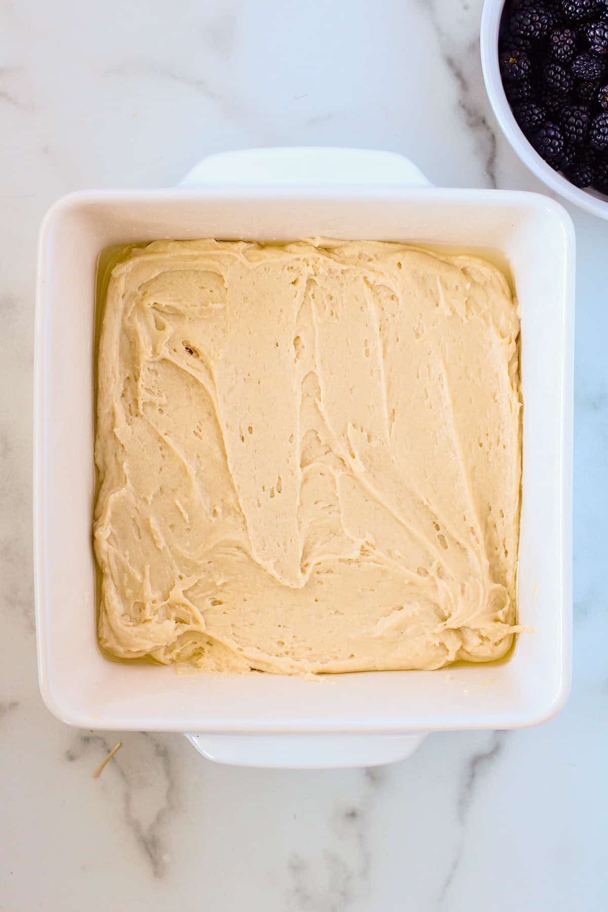 A square white baking dish with an even layer of unbaked blackberry cobbler batter sits on a marble surface; a bowl of blackberries is partially visible in the corner.