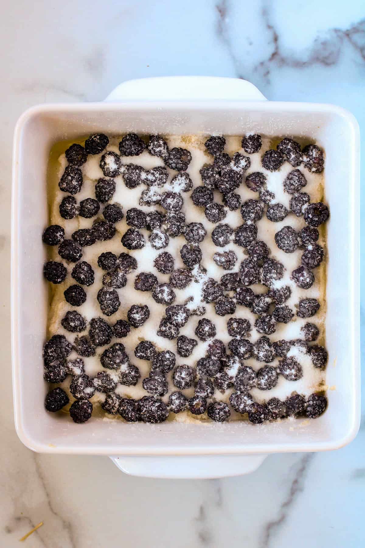 Square white baking dish with an unbaked blackberry cobbler, topped with scattered blackberries and granulated sugar on a marble surface.