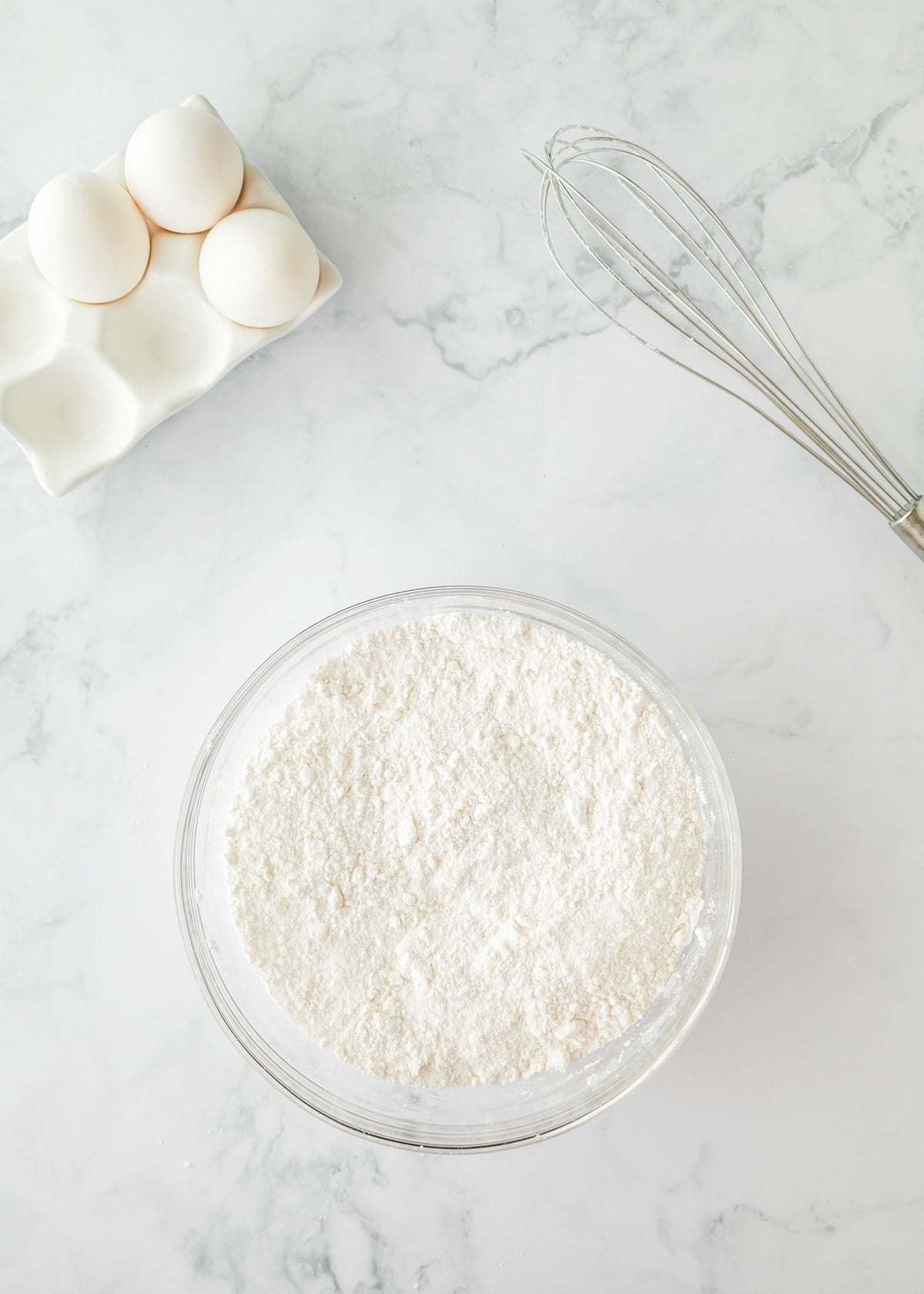A glass bowl of flour, three eggs in a ceramic holder, and a metal whisk on a white marble countertop—everything you need to start baking the perfect yellow cake.