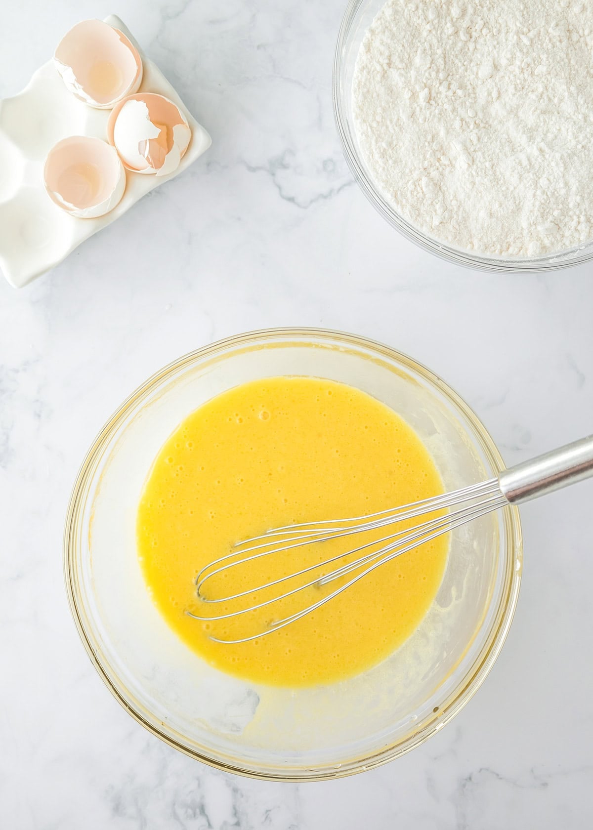 A bowl of beaten eggs with a whisk, next to a bowl of flour and cracked eggshells on a marble countertop—ingredients coming together for the perfect yellow cake.