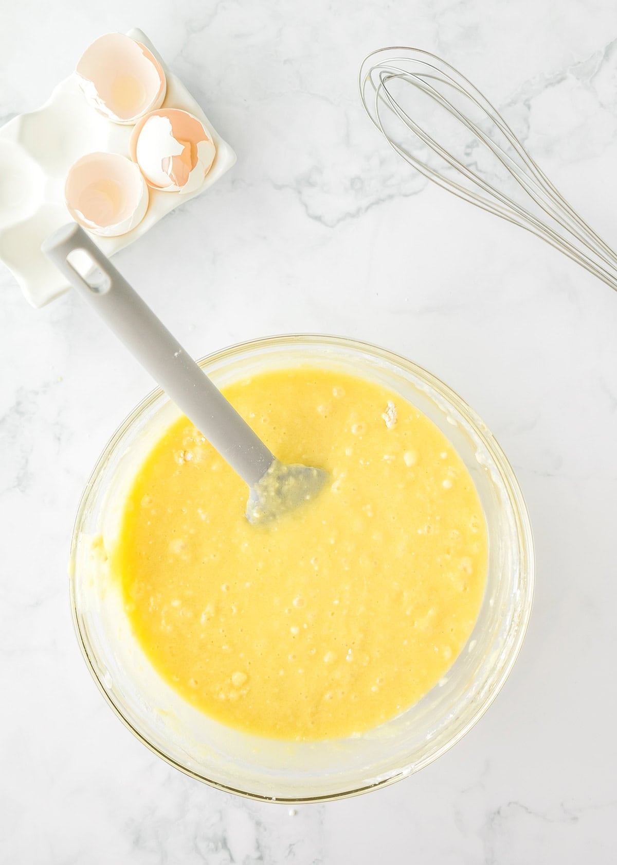 A glass bowl of yellow cake batter with a spatula, next to cracked eggshells and a whisk on a white marble surface.