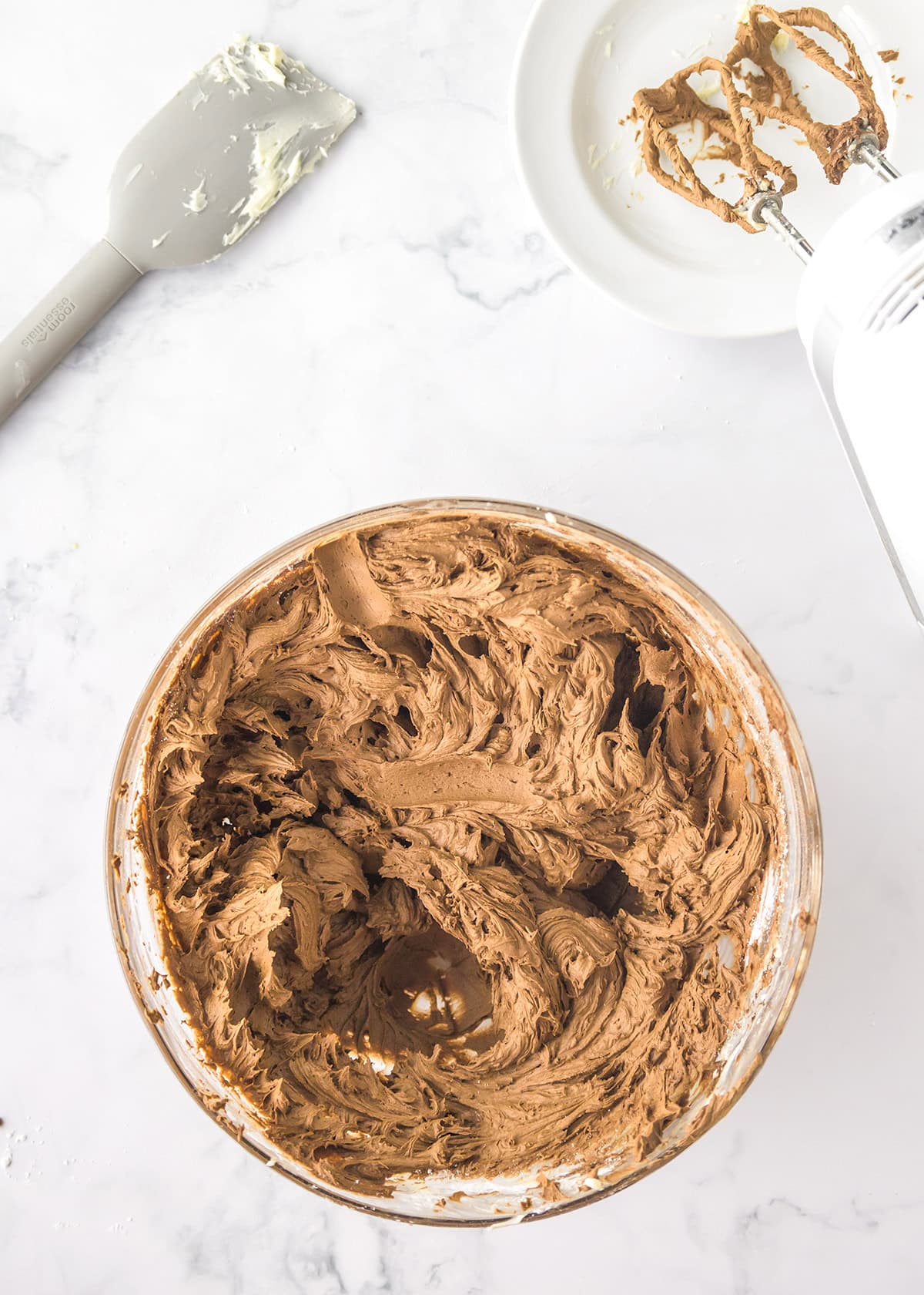 A glass bowl of chocolate frosting with a spatula and a mixer on a white surface, ready to be spread onto a freshly baked yellow cake.