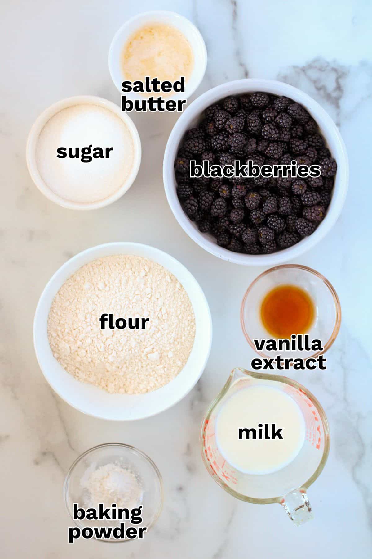 Seven labeled bowls containing salted butter, sugar, blackberries, flour, vanilla extract, milk, and baking powder are arranged on a white marble surface—everything you need to create a classic blackberry cobbler.