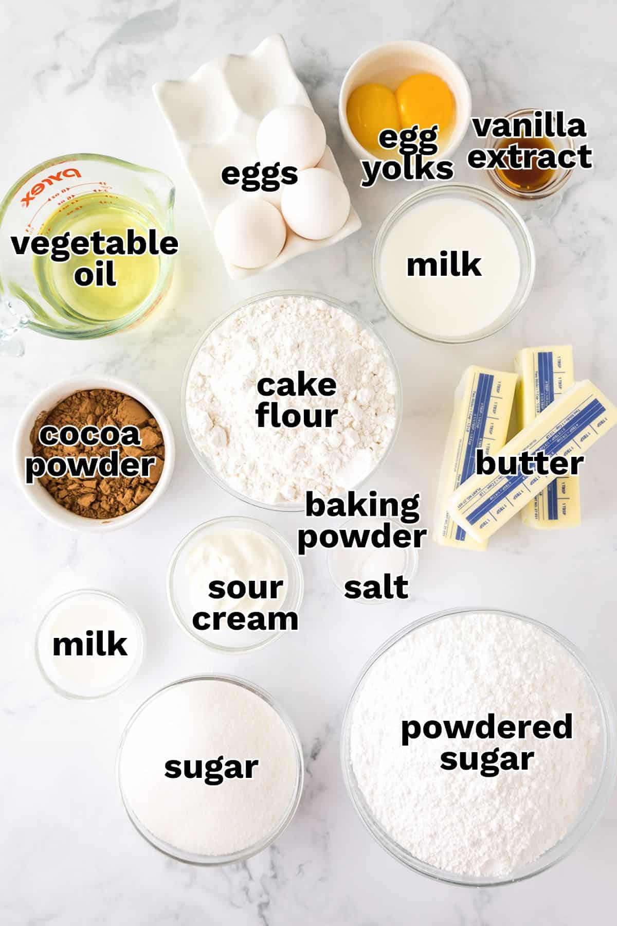 Various baking ingredients arranged on a white surface, including eggs, egg yolks, vanilla extract, milk, vegetable oil, cocoa powder, cake flour, baking powder, salt, sour cream, butter, sugar, and powdered sugar—everything you need for the perfect Yellow Cake.