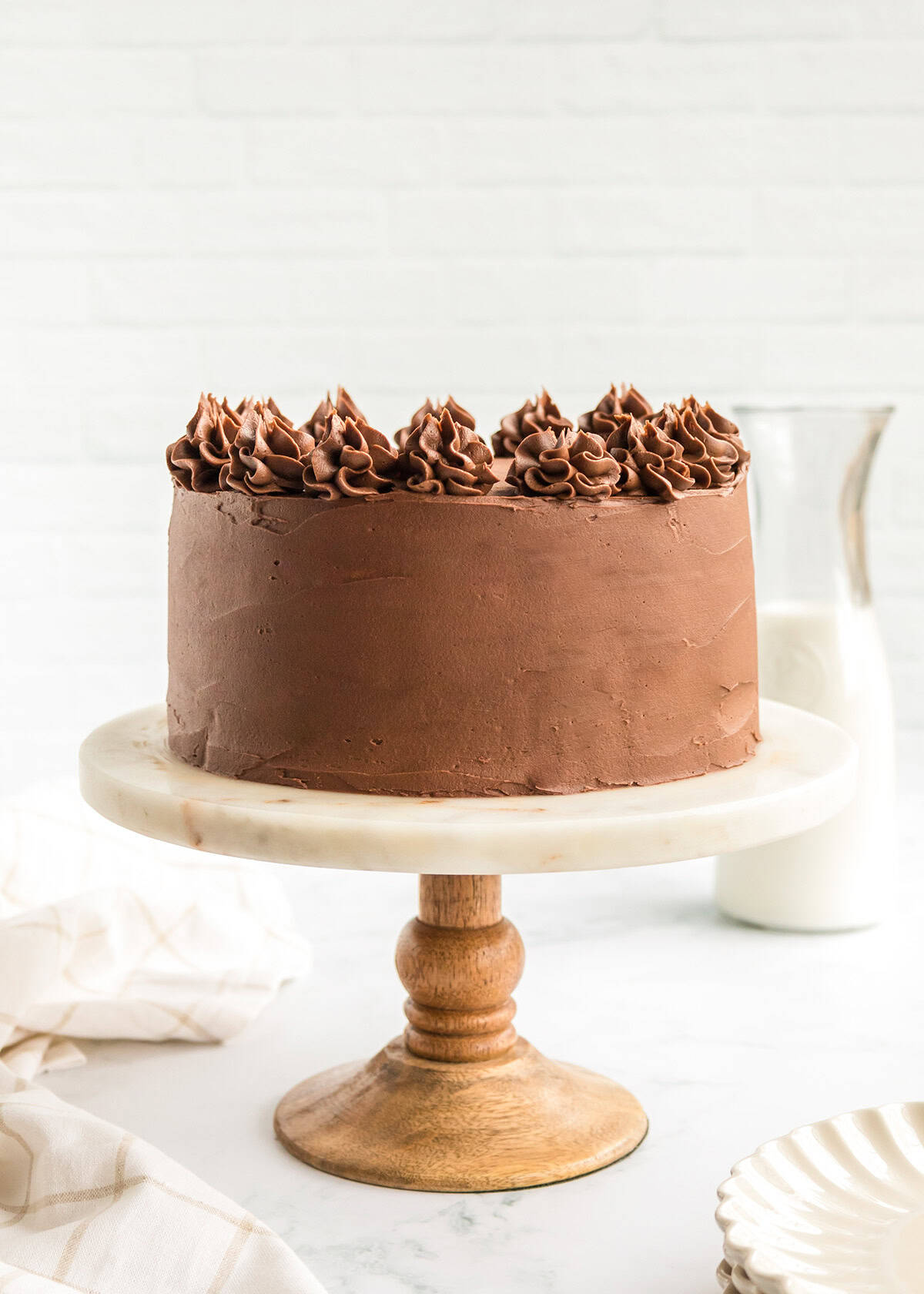 Chocolate frosted yellow cake with piped decorations on a wooden cake stand, accompanied by a glass of milk and a cloth napkin nearby.