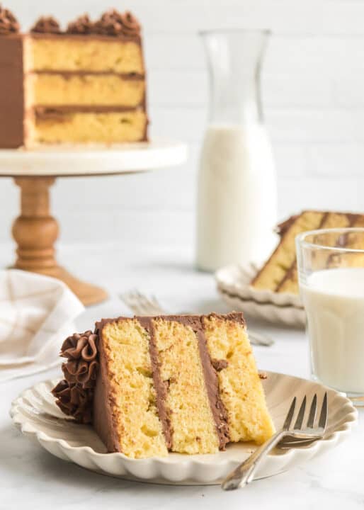 A slice of classic Yellow Cake with three layers and rich chocolate frosting sits on a plate with a fork, next to a glass of milk. The rest of the Yellow Cake and a milk carafe are in the background, creating a cozy scene perfect for any dessert lover.