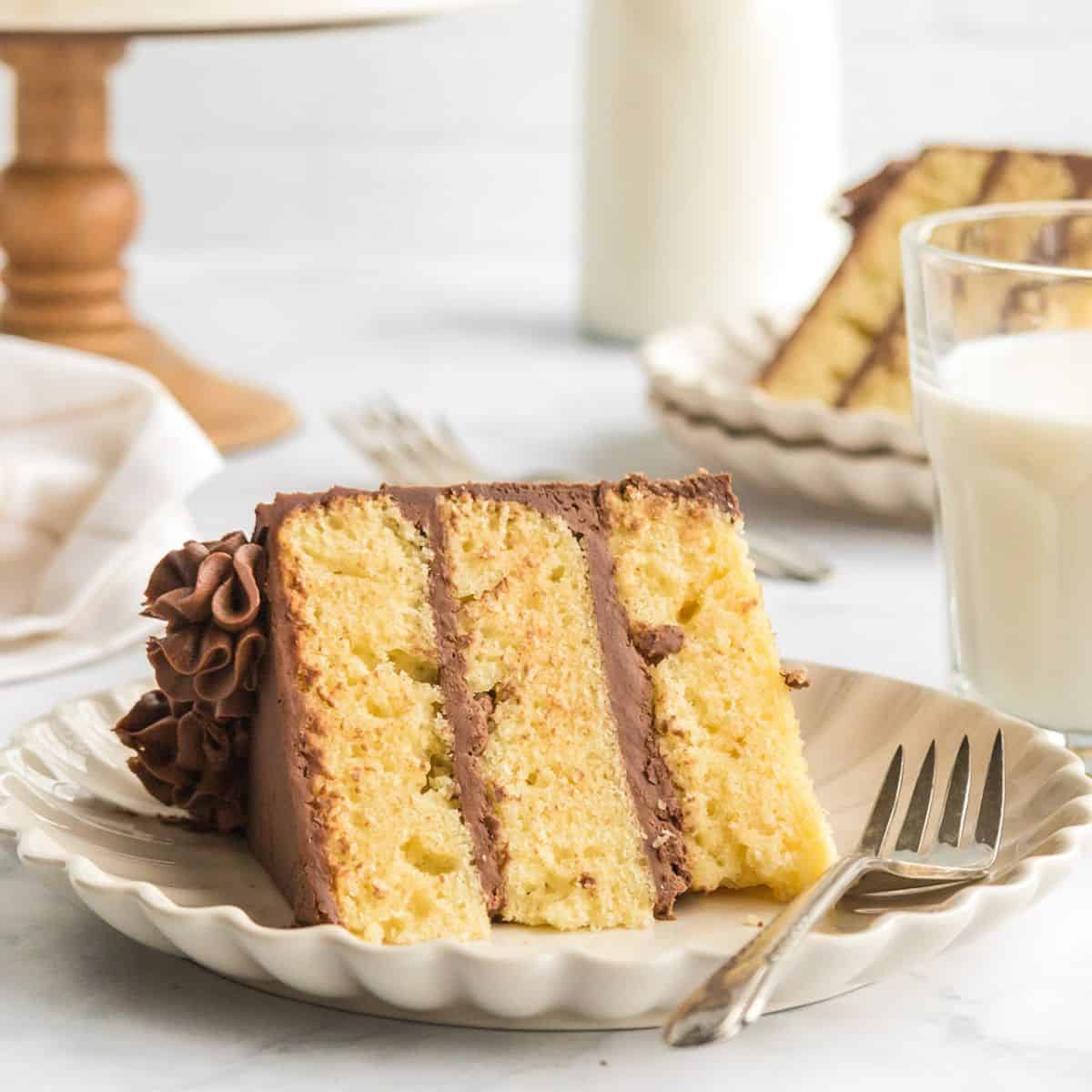 A slice of Yellow Cake with chocolate frosting sits on a plate with a fork, next to a glass of milk. Another slice and a bottle of milk are in the background.