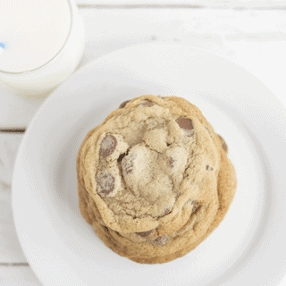 A stack of thin and chewy chocolate chip cookies on a white plate with a glass of milk.
