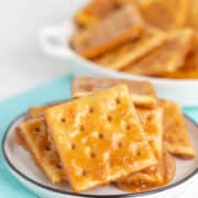 A close-up of caramel-coated saltine cracker toffee stacked on a small plate with more in a dish behind, set on a light blue napkin.