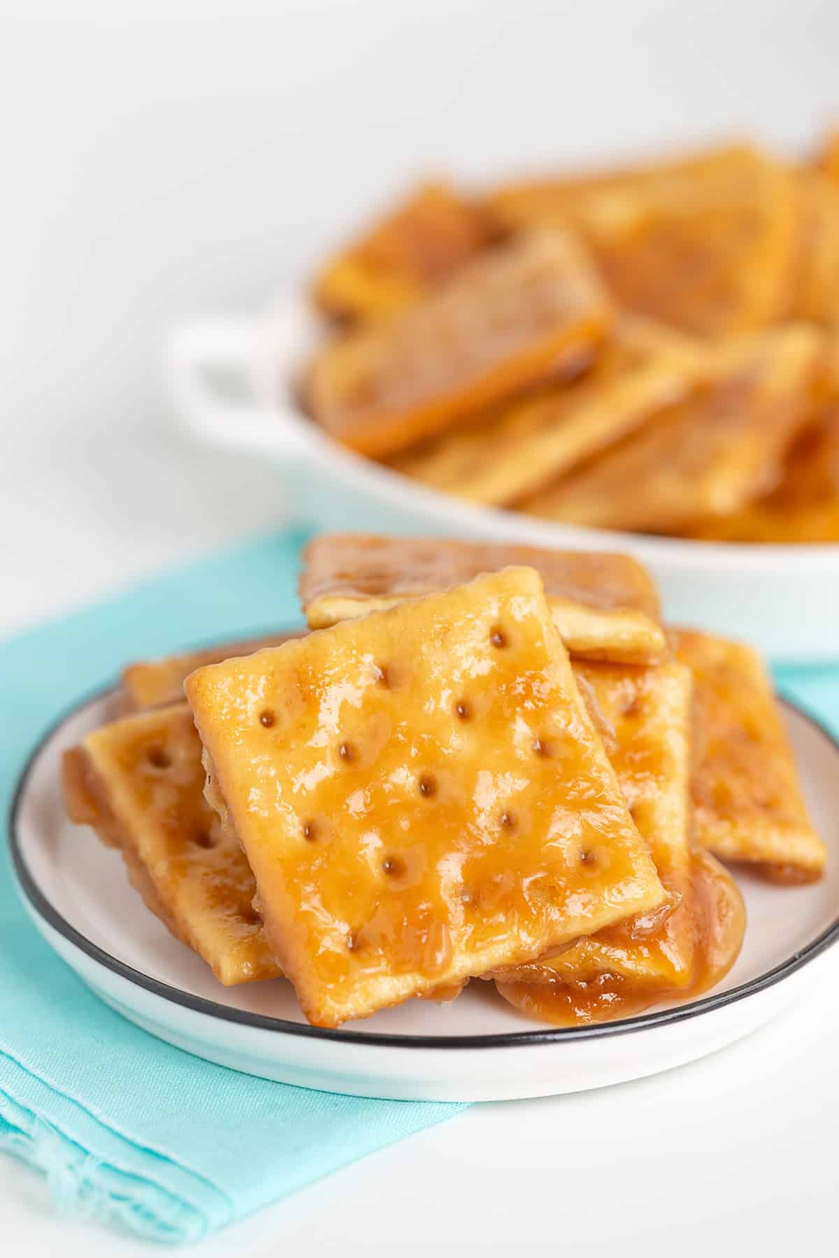 A close-up of caramel-coated saltine cracker toffee stacked on a small plate with more in a dish behind, set on a light blue napkin.
