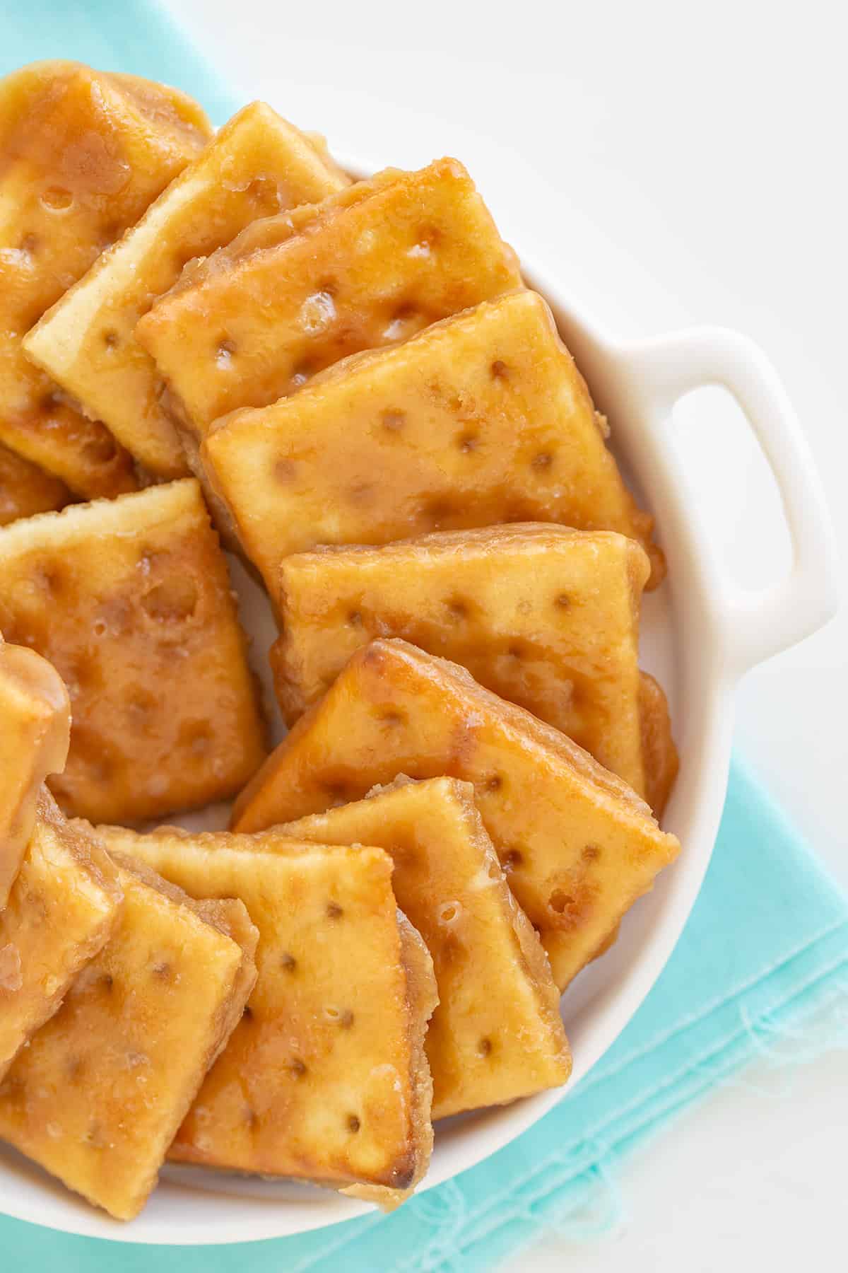 A bowl brimming with Saltine Cracker Toffee, featuring caramel-coated squares, sits elegantly on a turquoise cloth.