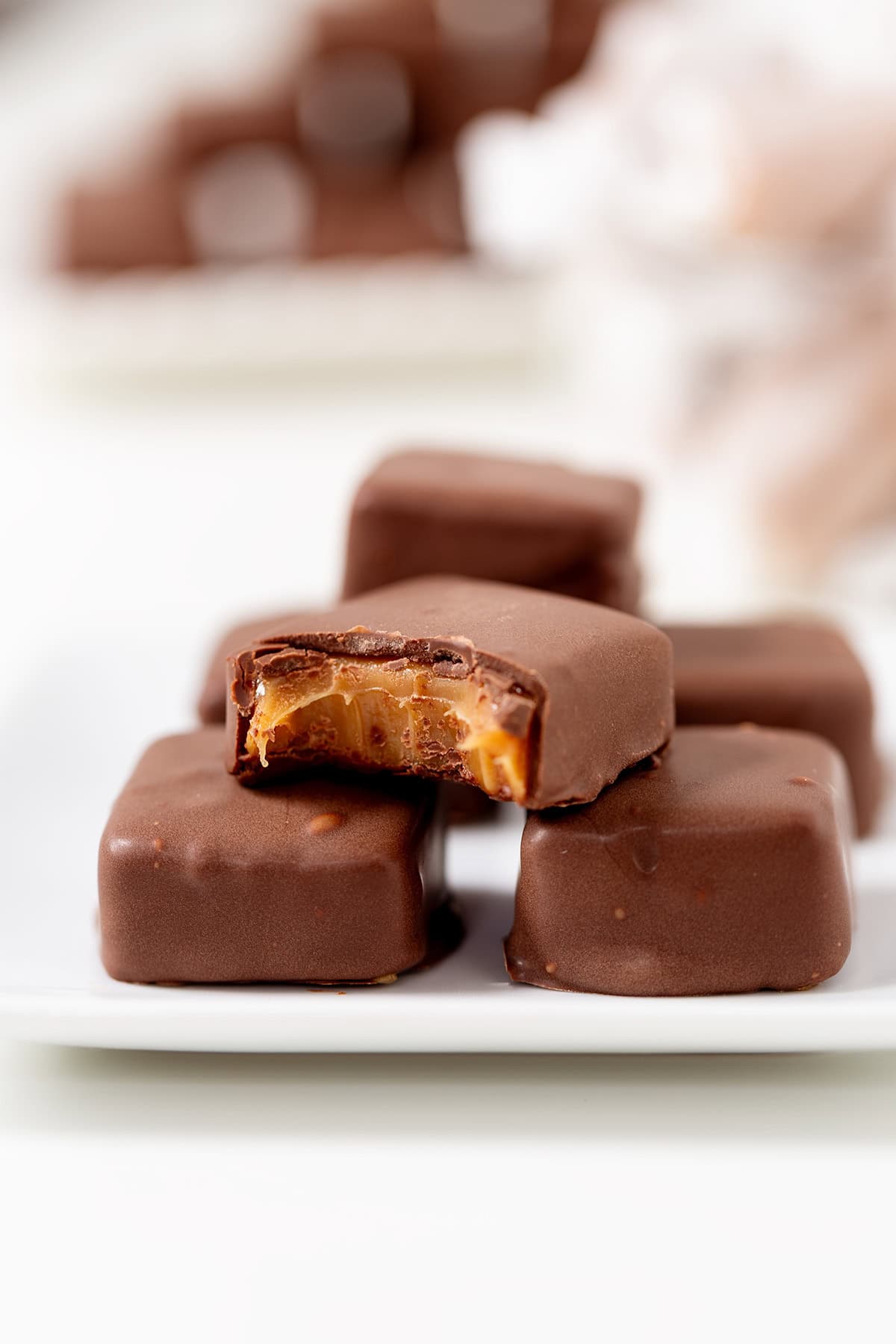 A close-up of Soft & Chewy Caramels coated in chocolate on a white plate, with one candy showing a bite that reveals the gooey caramel filling inside.