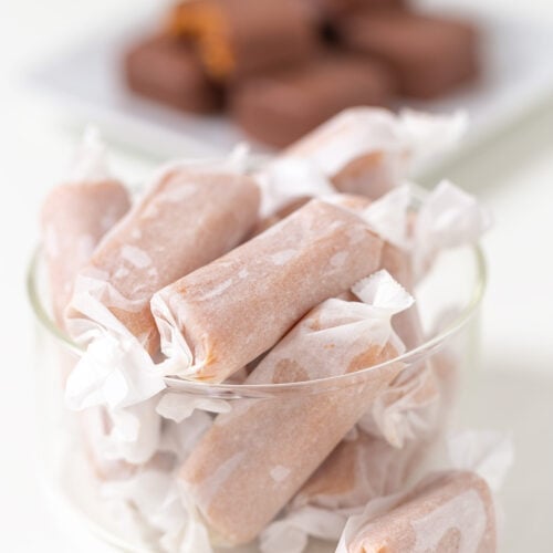 A glass bowl filled with Soft & Chewy Caramels, with a plate of chocolate-covered candies blurred in the background.