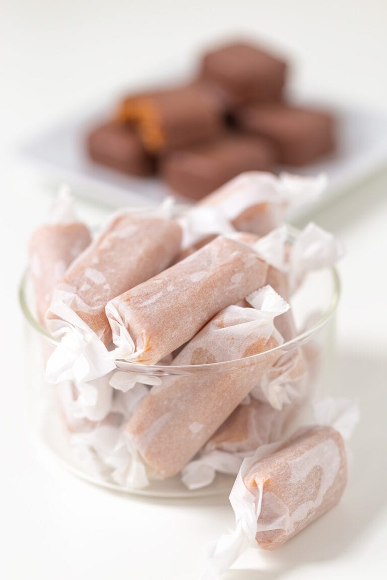 A glass bowl filled with Soft & Chewy Caramels, with a plate of chocolate-covered candies blurred in the background.