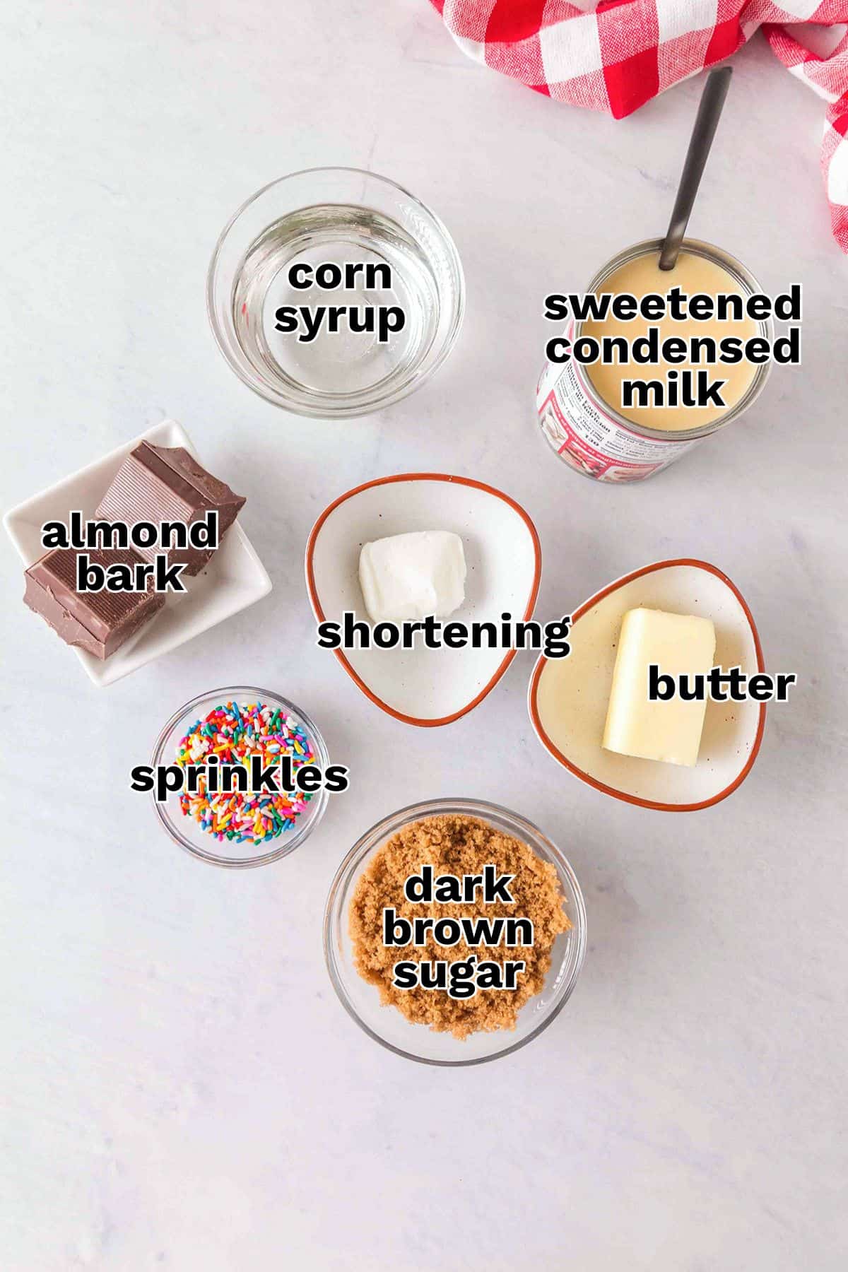 Various baking ingredients are arranged on a white surface, each labeled: corn syrup, sweetened condensed milk, almond bark, shortening, butter, sprinkles, and dark brown sugar&mdash;perfect for making Soft & Chewy Caramels.