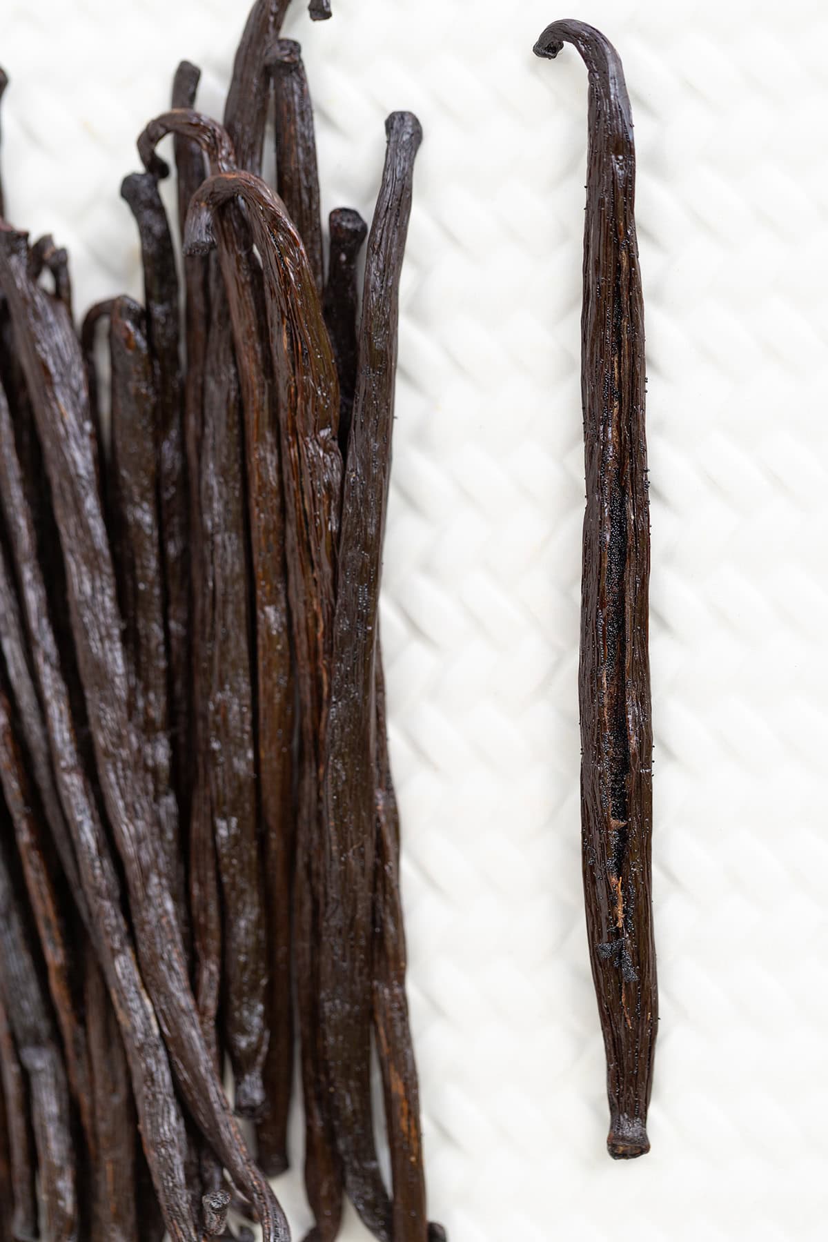 Several dark brown vanilla beans, ideal for making homemade vanilla extract, are arranged on a textured white surface, with one vanilla bean separated from the group.