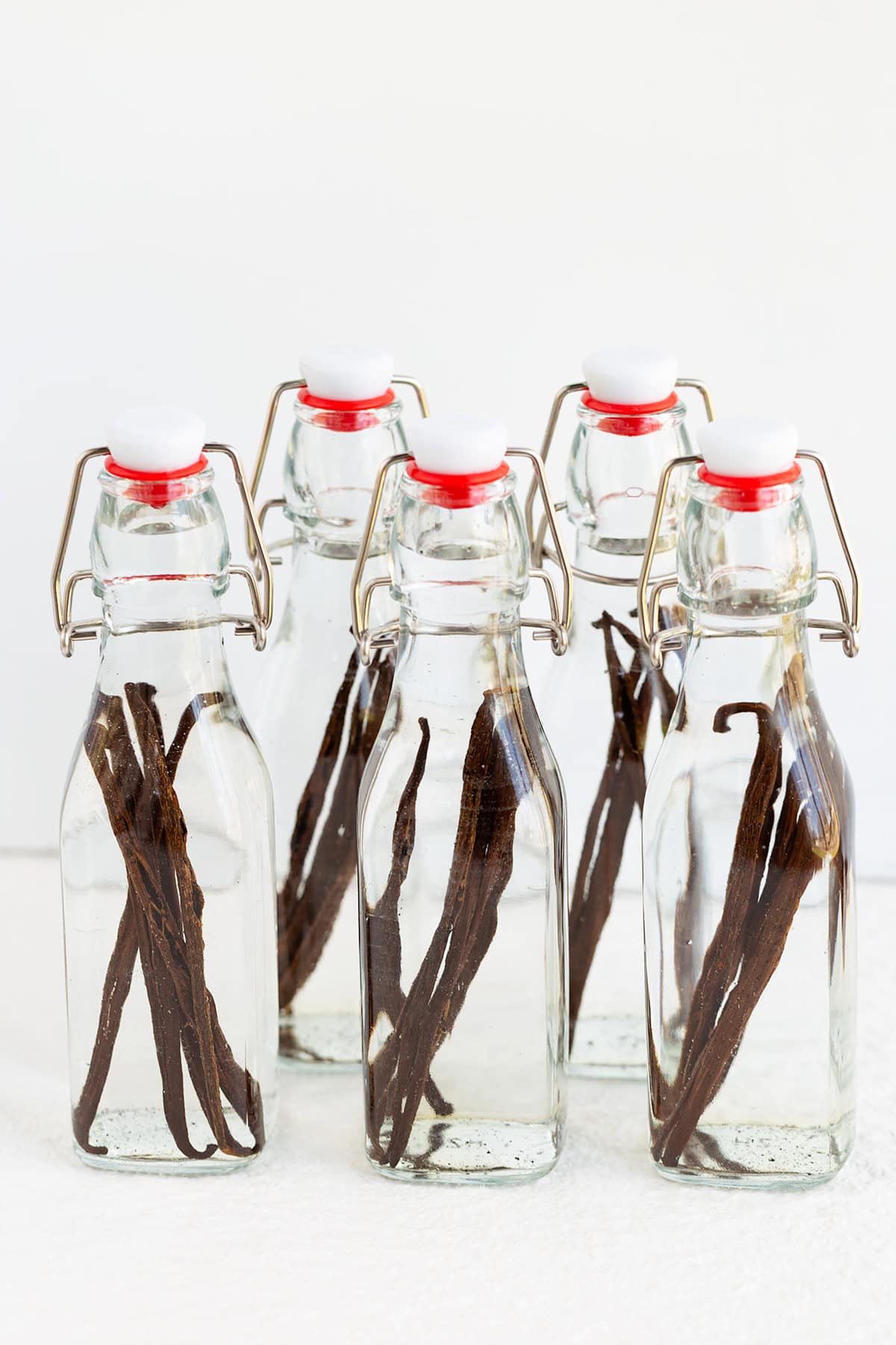 Five clear glass bottles with metal clasps, each containing vanilla beans soaking in a transparent liquid, are arranged on a white surface&mdash;perfect for creating rich homemade vanilla extract.