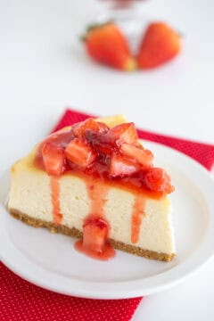 A slice of New York Style Cheesecake topped with strawberry sauce on a white plate, set on a red polka-dot napkin, with two blurred strawberries in the background.