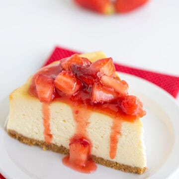A slice of New York Style Cheesecake topped with strawberry sauce on a white plate, set on a red polka-dot napkin, with two blurred strawberries in the background.