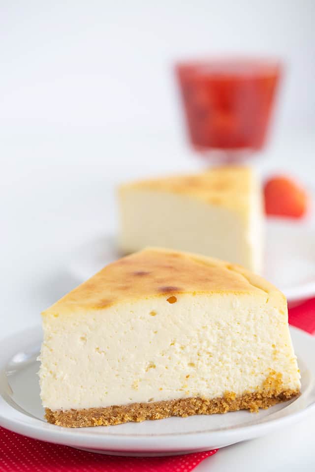 A slice of New York Style Cheesecake sits on a white plate with another slice and a red drink in the background.