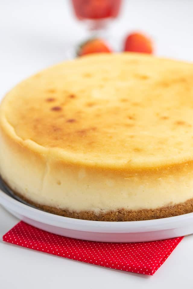 A whole New York Style Cheesecake on a white plate with a red napkin underneath. In the background, there is a glass with strawberries.