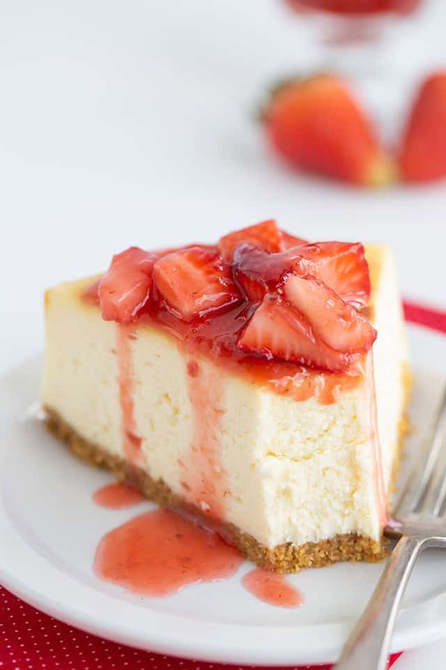 A slice of New York Style Cheesecake topped with strawberry sauce and sliced strawberries on a white plate, with a fork beside it.
