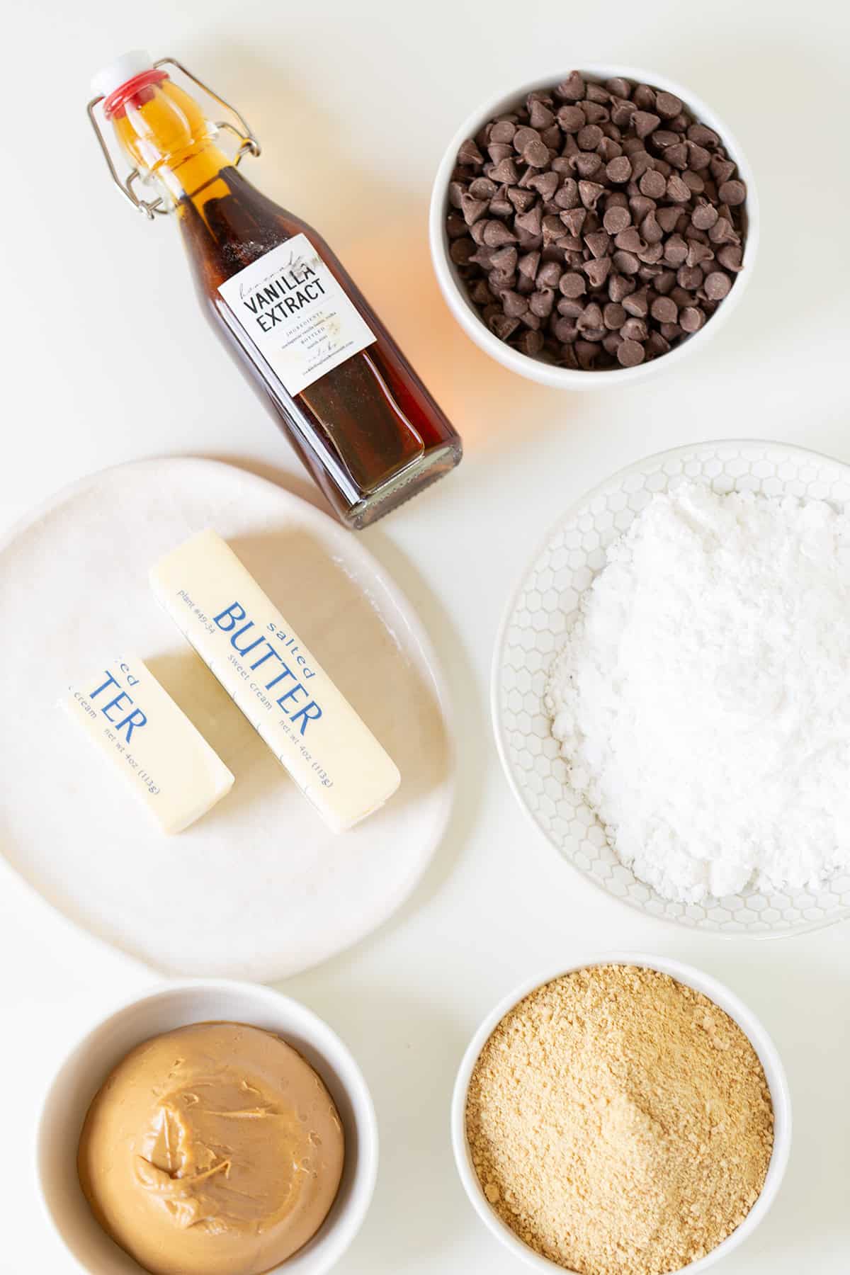 Ingredients for Peanut Butter Bars on a white surface: vanilla extract bottle, chocolate chips, powdered sugar, butter sticks on a plate, peanut butter in a bowl, and crushed graham crackers in a bowl.
