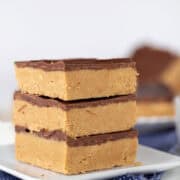Three luscious peanut butter bars with a chocolate topping are stacked on a white plate, with a blue patterned cloth beneath. More of these delectable peanut butter bars are blurred in the background.