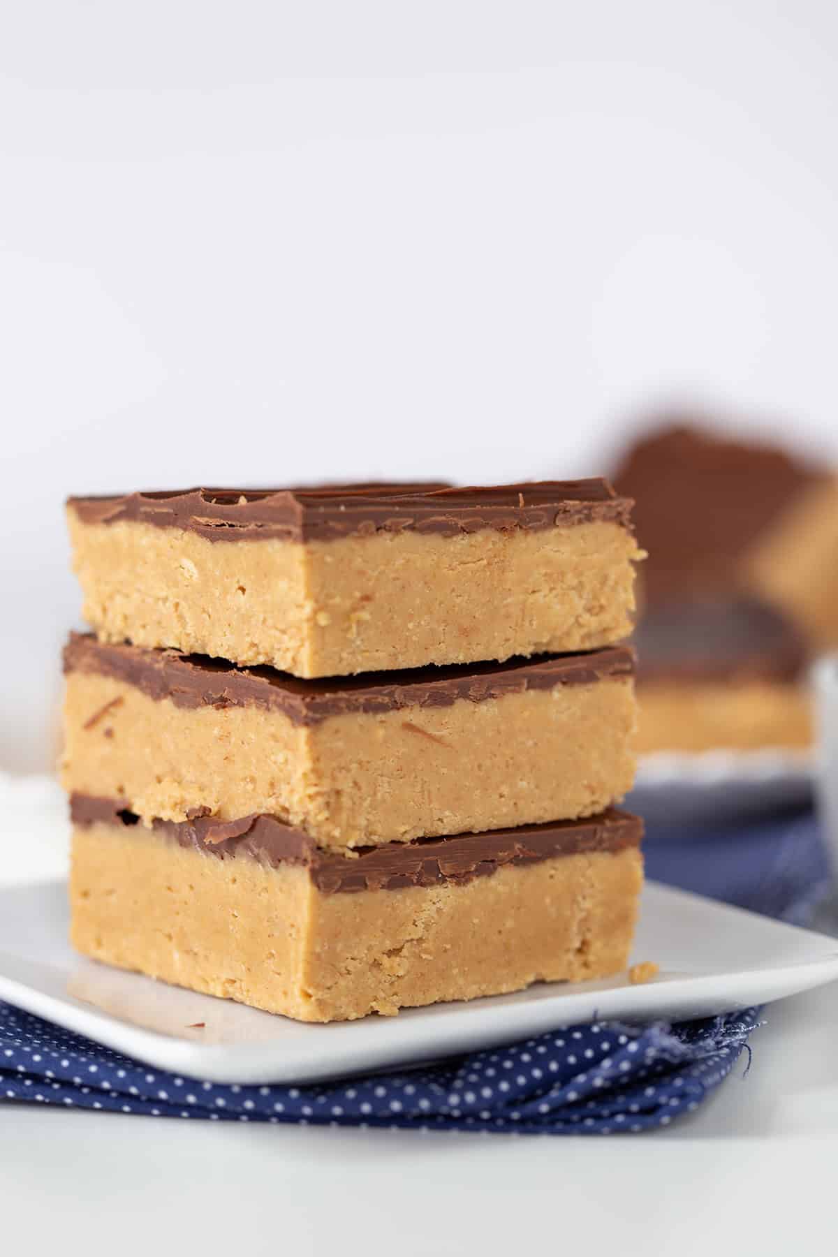 Three luscious peanut butter bars with a chocolate topping are stacked on a white plate, with a blue patterned cloth beneath. More of these delectable peanut butter bars are blurred in the background.