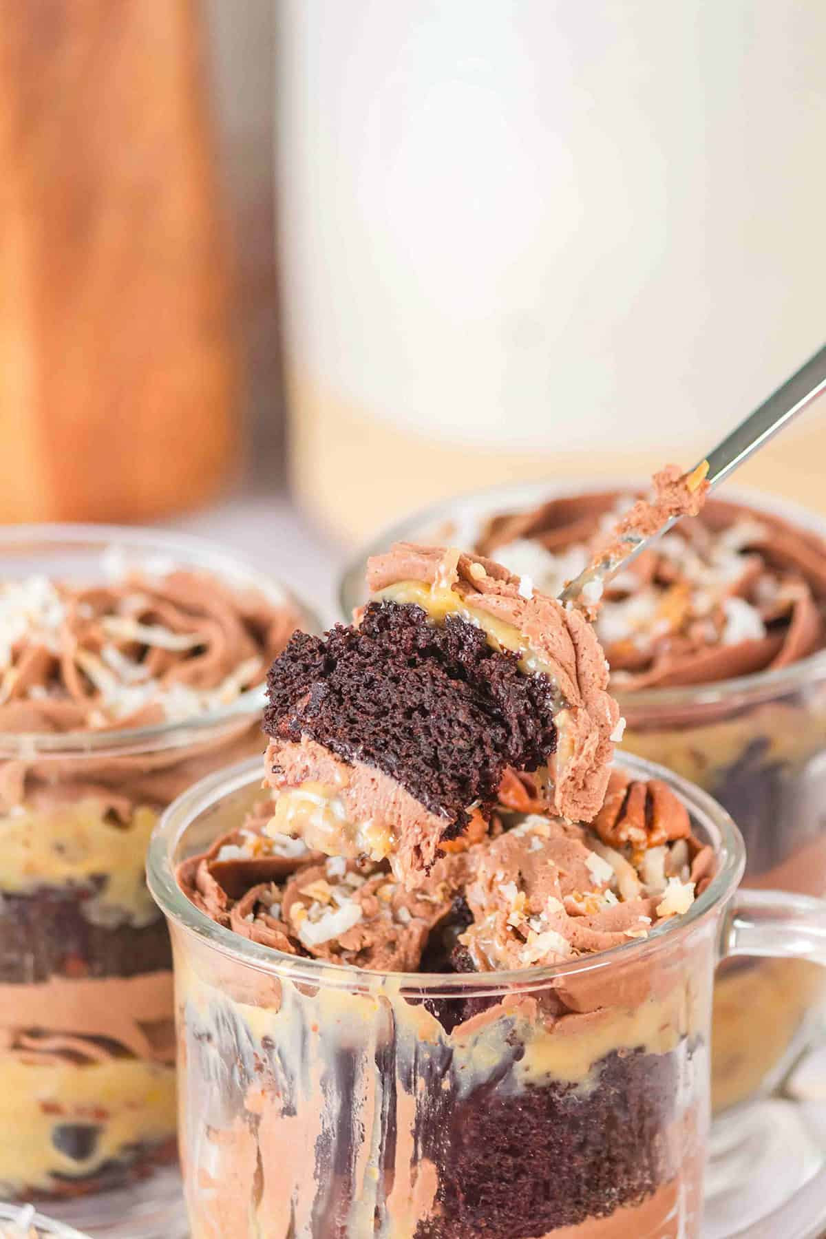 A close-up of a German Chocolate Trifle in a glass cup reveals chocolate and caramel layers, with a spoon lifting a piece. It's beautifully topped with shredded coconut.
