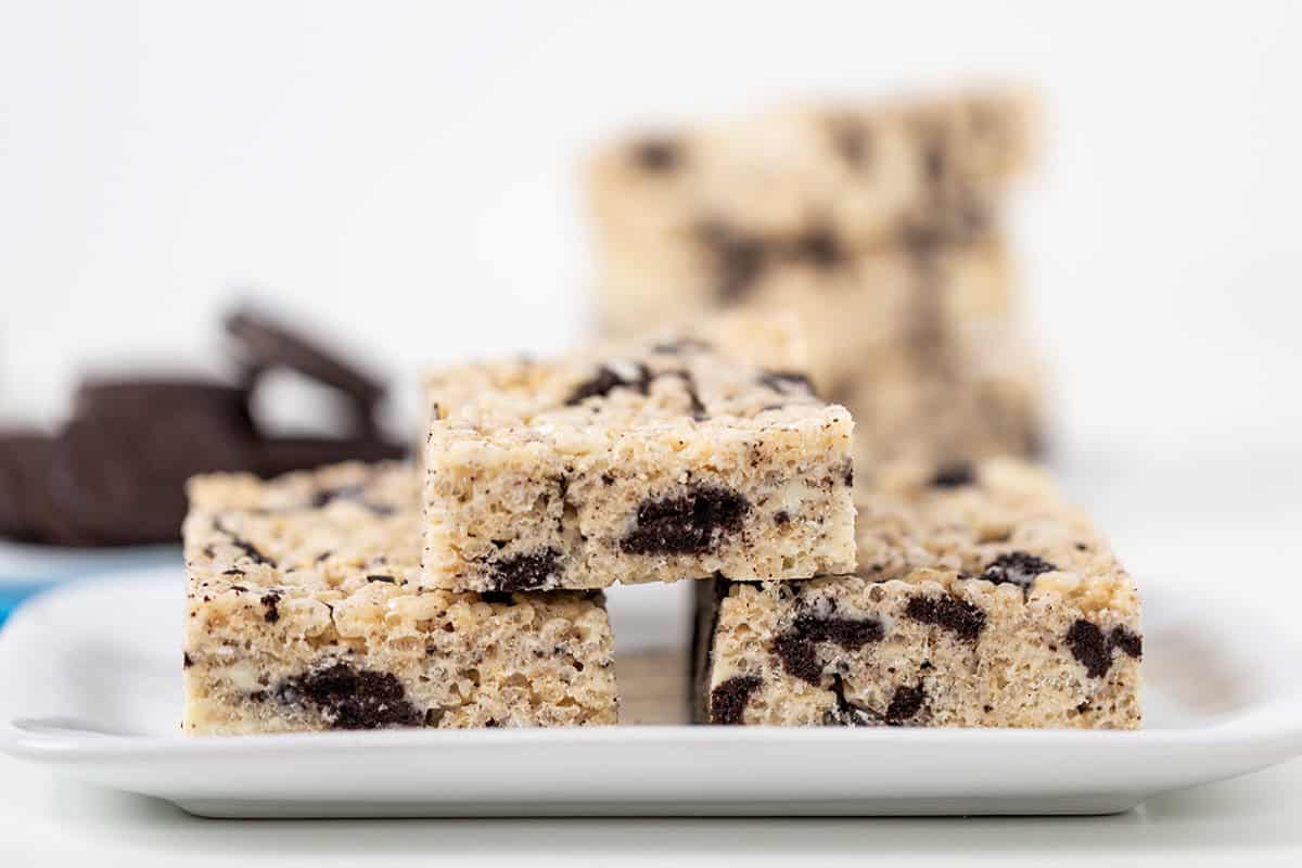 Three Cookies & Cream Rice Crispy Treats with visible chocolate cookie pieces are stacked on a white plate, with more treats and cookies blurred in the background.