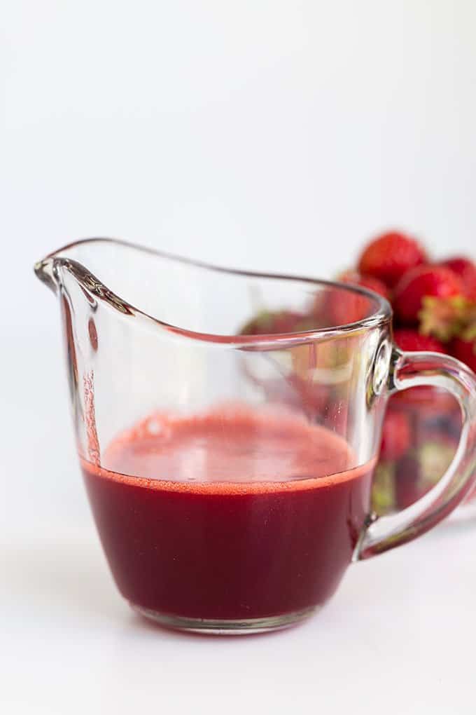 serving glass with homemade strawberry syrup in it and fresh strawberries behind it on a white background