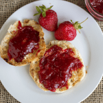 A white plate with two English muffin halves spread with strawberry rhubarb jam, two fresh strawberries, and a jar of jam on a burlap mat.