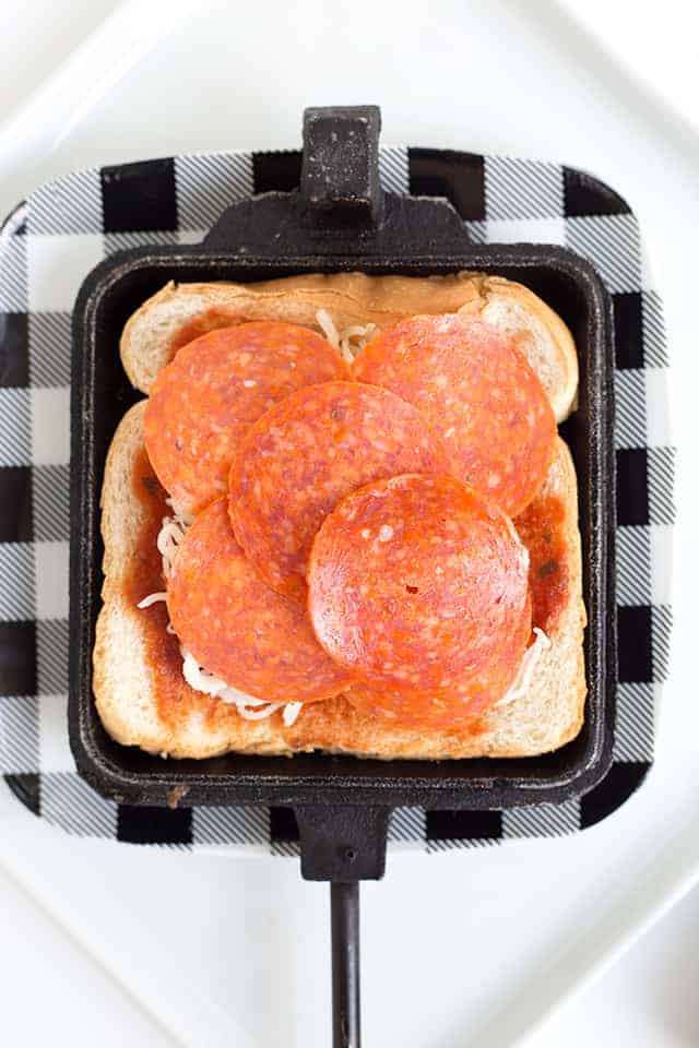Pizza Mountain Pies - These delicious pizza mountain pies are the perfect treat while camping! These pizza mountain pies are made over the campfire coals with a pie iron. They take minutes to make and are the most amazing pizza pies you'll have! Enjoy the crunchy, buttery outside and the gooey cheese and pepperoni pizza inside. #cookiedoughandovenmitt #camping #campingrecipes #pizza adding pepperoni to a pizza mountain pie maker