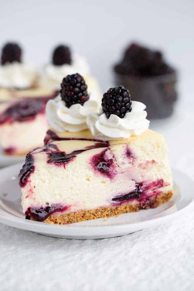 Slice of blackberry cheesecake on white plate with blackberries in background