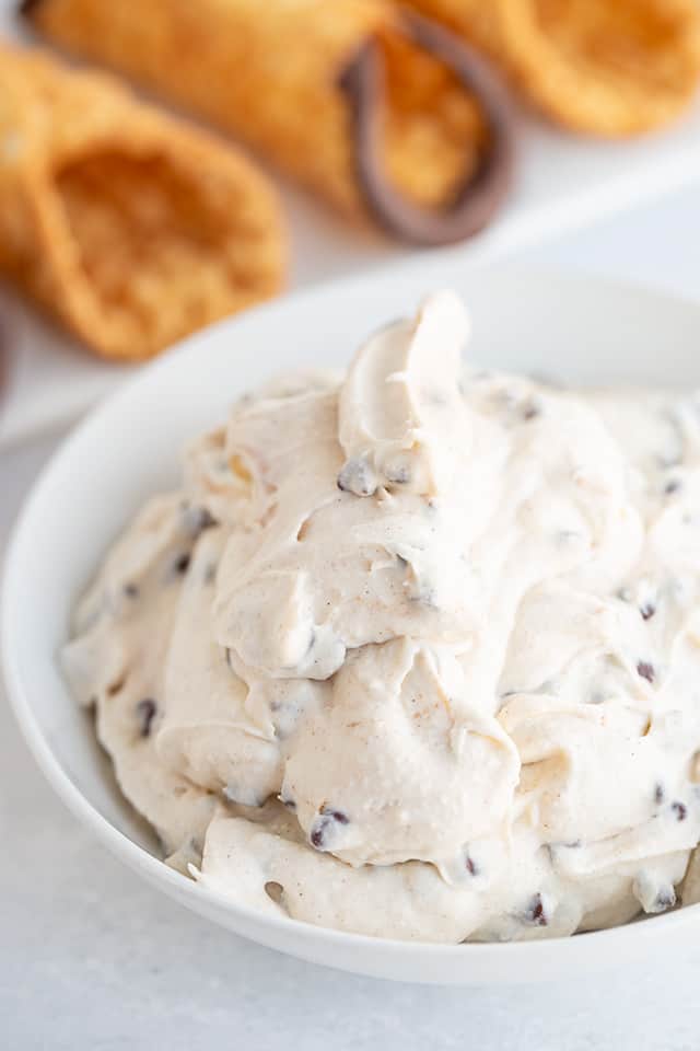 a bowl of cannoli cream filling with shells on a platter behind it