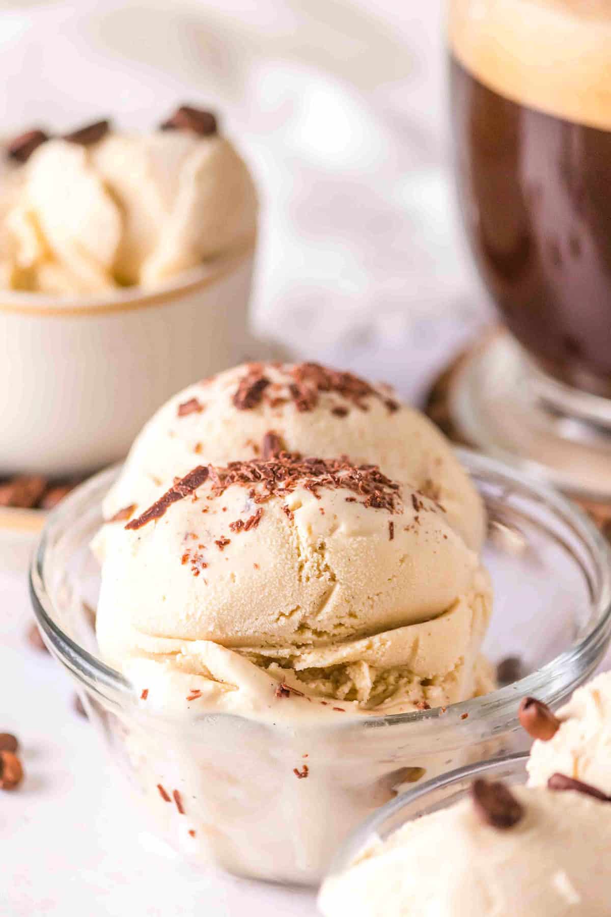 A bowl of vanilla ice cream topped with chocolate shavings sits front and center, while another bowl, this one filled with rich coffee ice cream, and a glass of cold beverage linger enticingly in the background.