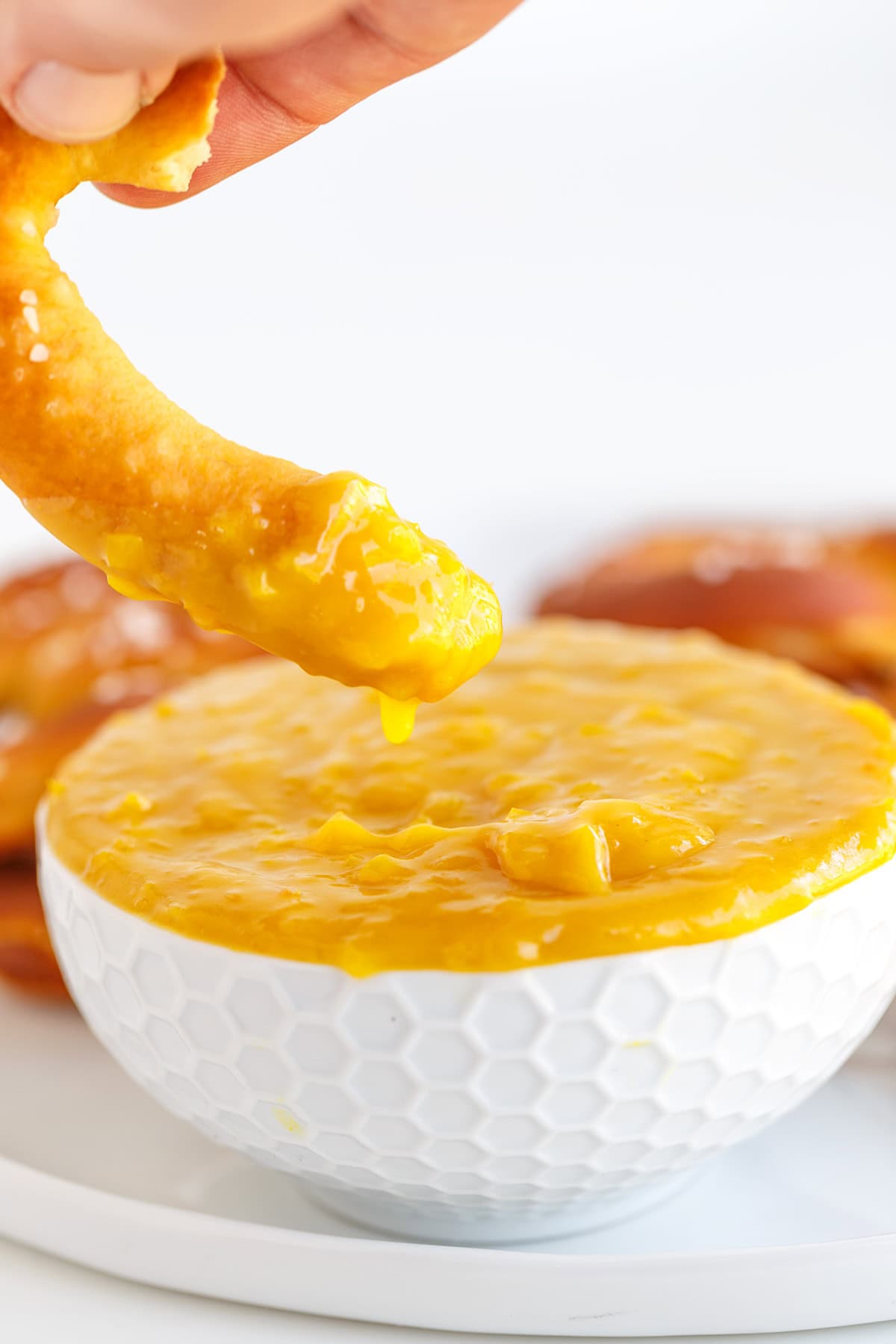 A hand dips a pretzel into a white bowl filled with yellow cheese sauce and a touch of Hot Pepper Butter, with more pretzels visible in the background.