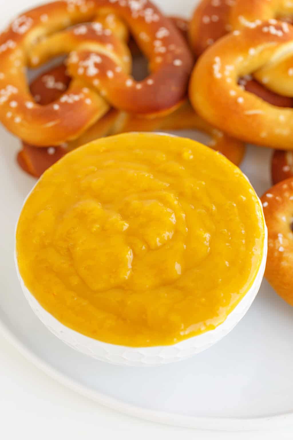 A white bowl filled with creamy yellow Hot Pepper Butter dipping sauce sits on a plate with several soft pretzels in the background.