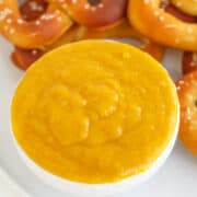A white bowl filled with creamy yellow Hot Pepper Butter dipping sauce sits on a plate with several soft pretzels in the background.