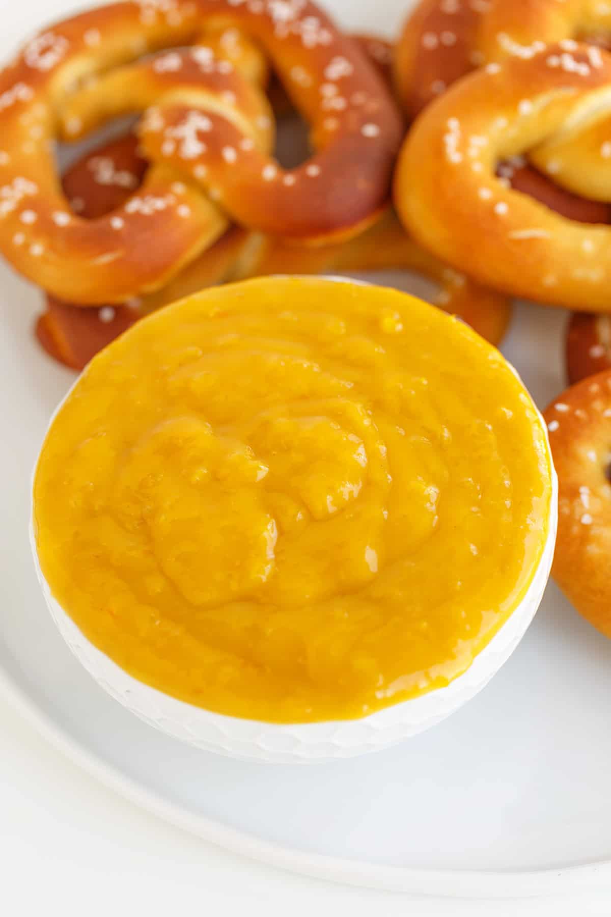 A white bowl filled with creamy yellow Hot Pepper Butter dipping sauce sits on a plate with several soft pretzels in the background.
