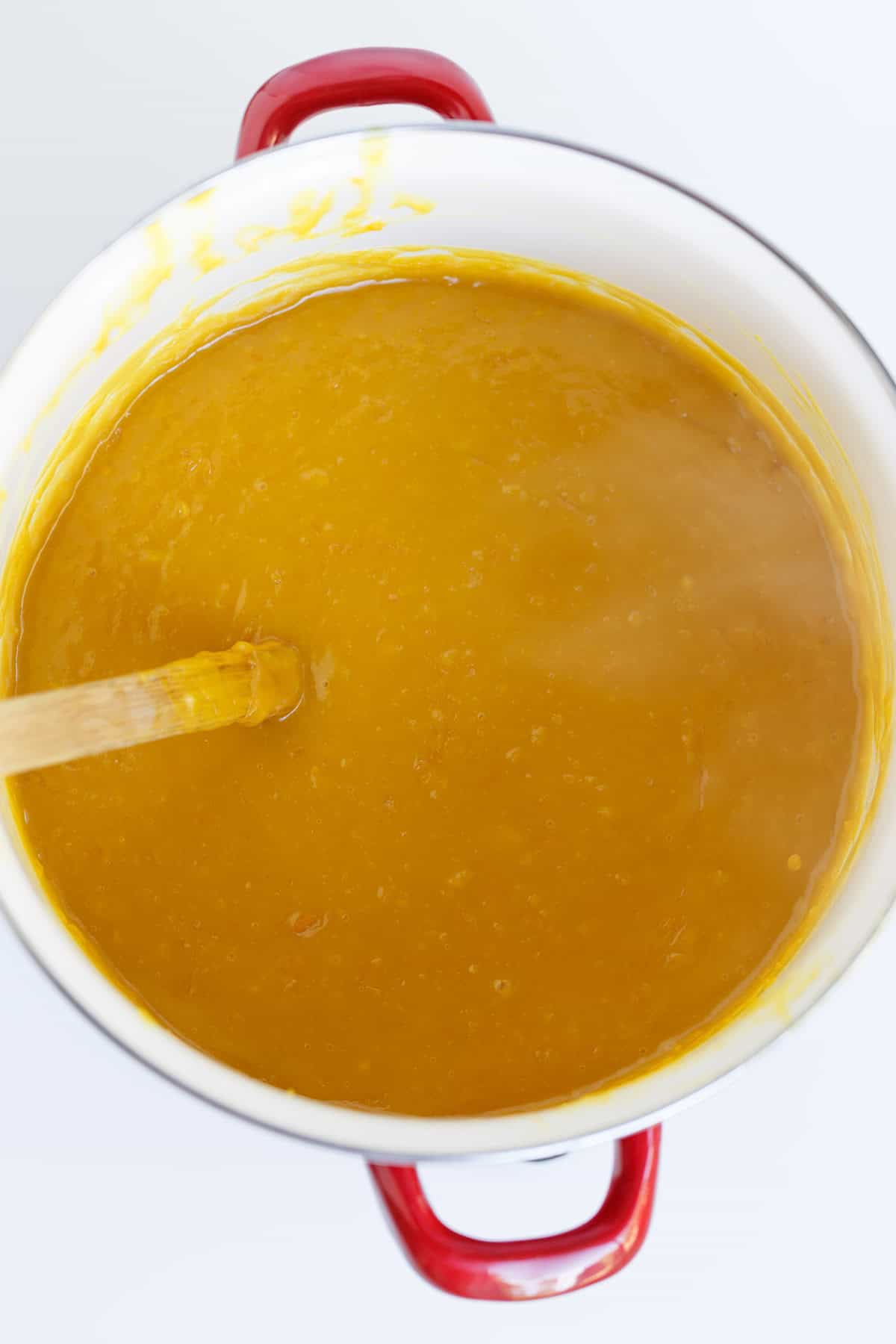 A white pot with red handles contains thick, yellow-orange soup being stirred with a wooden spoon, its rich aroma hinting at the addition of Hot Pepper Butter.