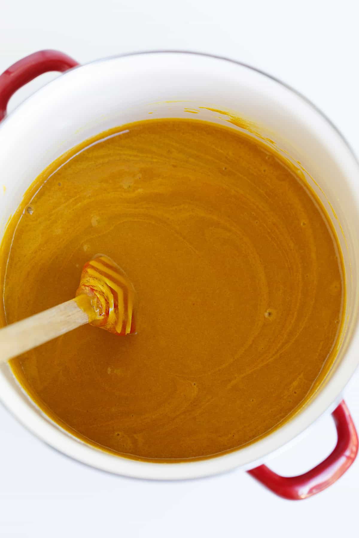 A pot with thick golden-brown Hot Pepper Butter being stirred by a wooden utensil, viewed from above on a white background.