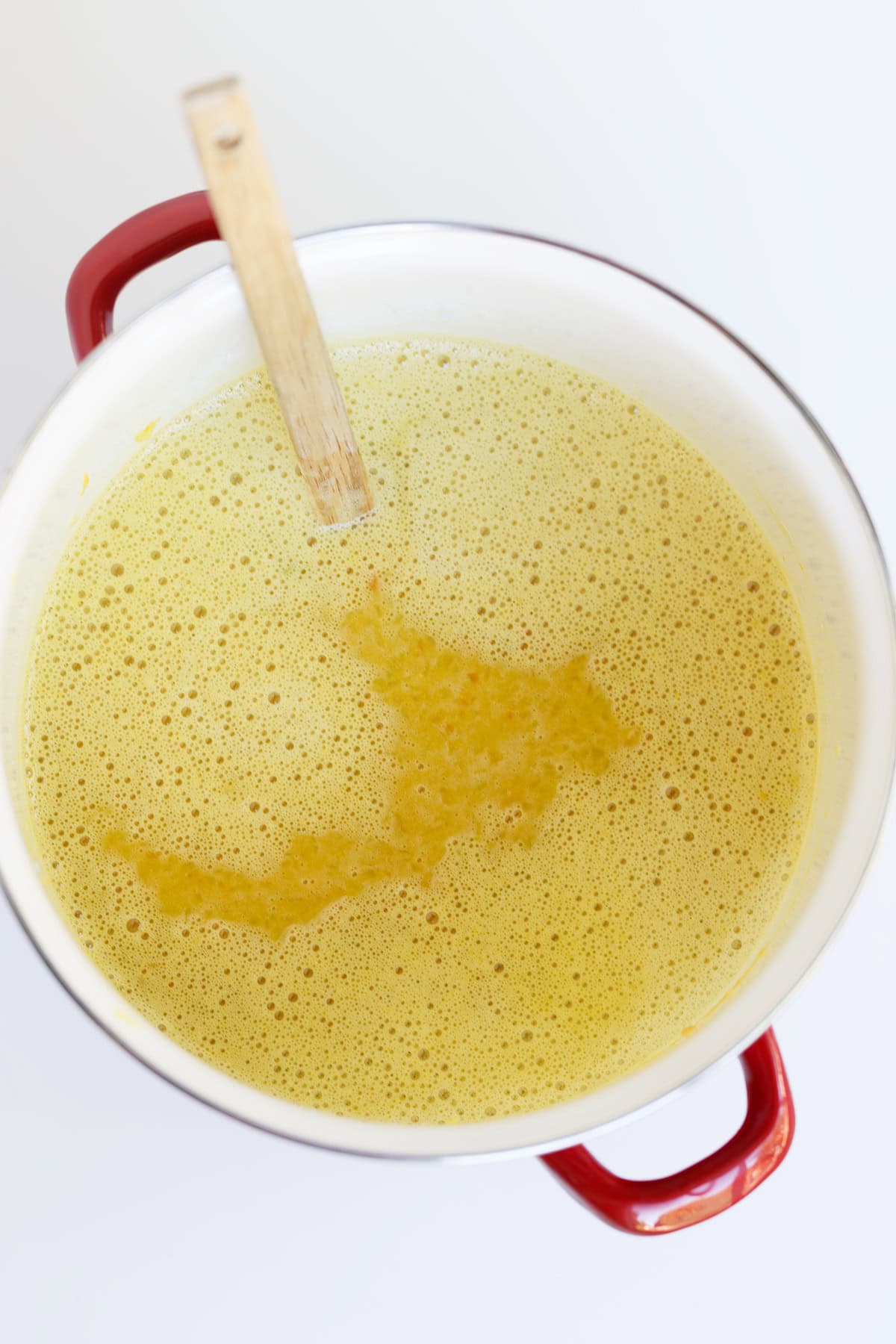 A white pot with red handles containing a yellow frothy Hot Pepper Butter and a wooden spoon resting inside, viewed from above.
