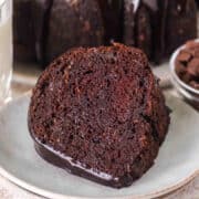 A slice of Triple Chocolate Zucchini Bundt Cake with rich chocolate glaze sits on a plate, accompanied by a glass of milk and a bowl of chocolate chips in the background.