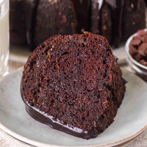 A slice of Triple Chocolate Zucchini Bundt Cake with rich chocolate glaze sits on a plate, accompanied by a glass of milk and a bowl of chocolate chips in the background.