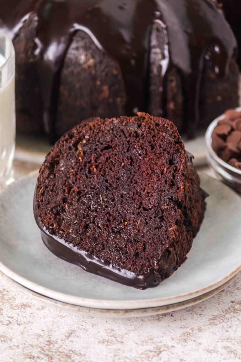 A slice of Triple Chocolate Zucchini Bundt Cake with rich chocolate glaze sits on a plate, accompanied by a glass of milk and a bowl of chocolate chips in the background.
